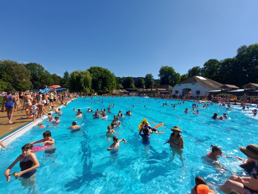Crowded Olympic-sized outdoor swimming pool at Guildford Lido filled with people on a bright, sunny summer day.