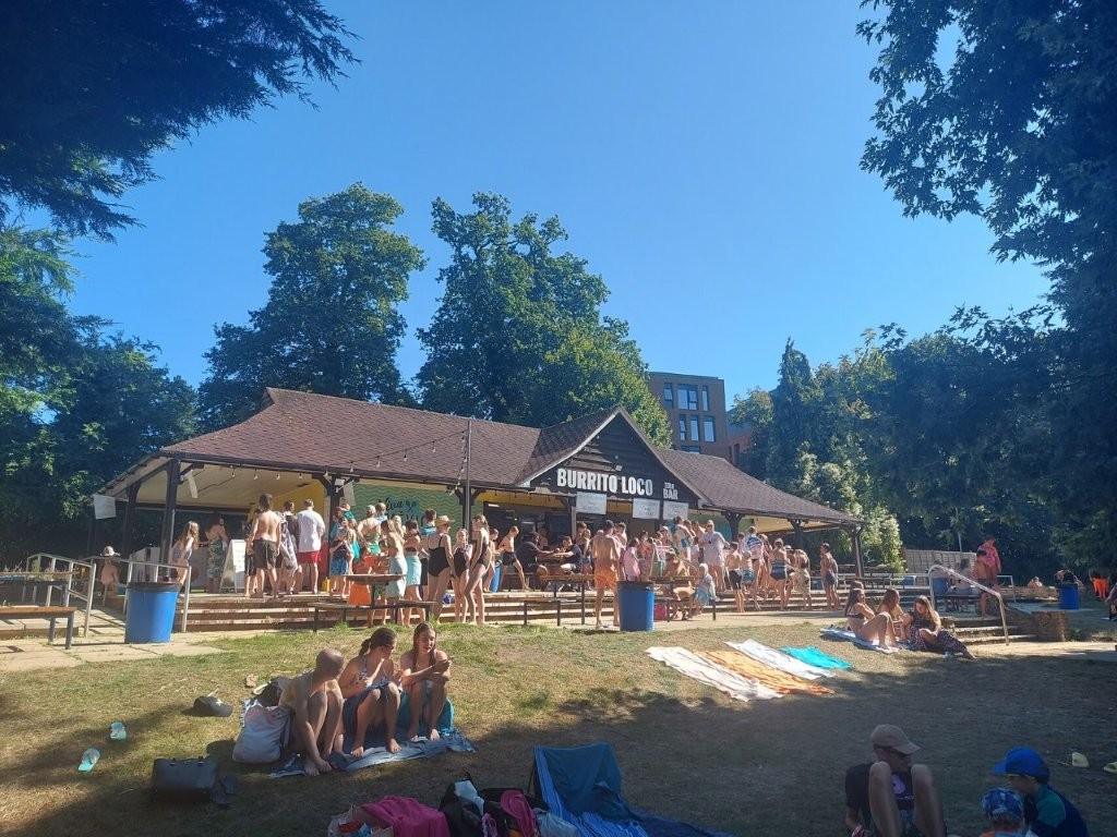 Crowds in swimwear outside the Burrito Loco food kiosk on the sunny grass lawns of Guildford Lido.