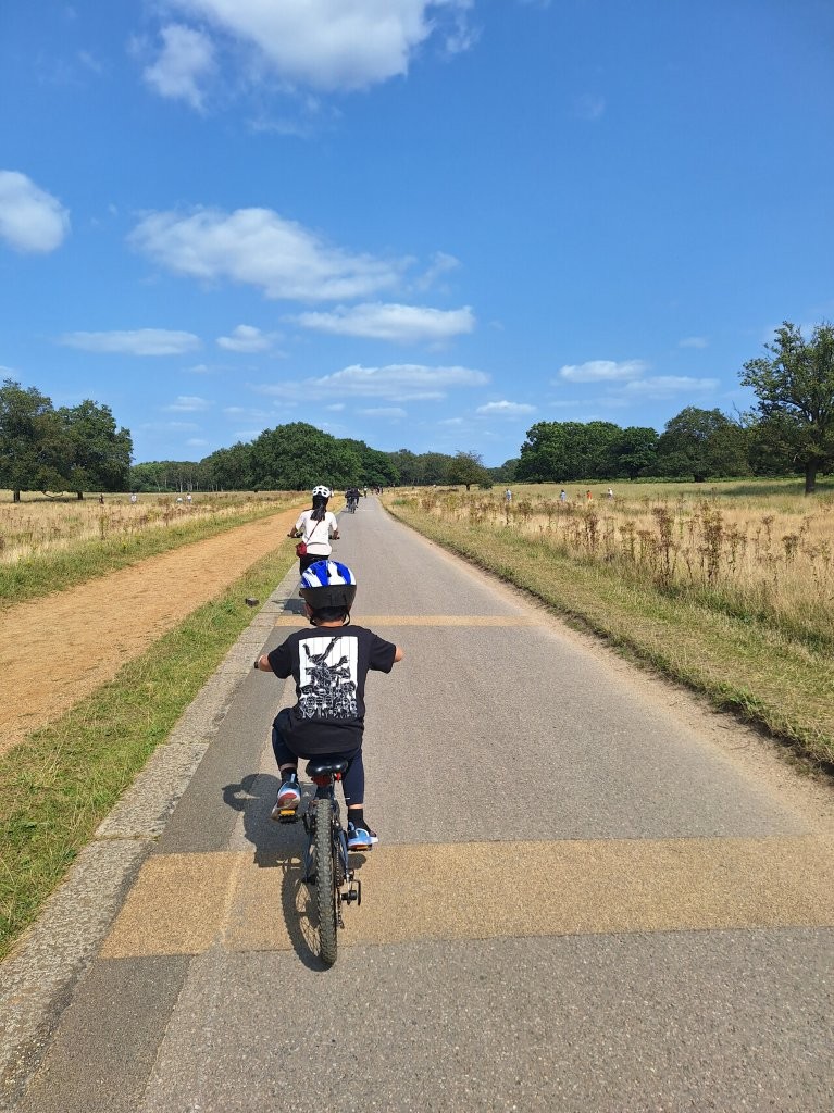 A child and adult cycling at richmond park on a paved path lined with dry grassy fields under a sunny sky.